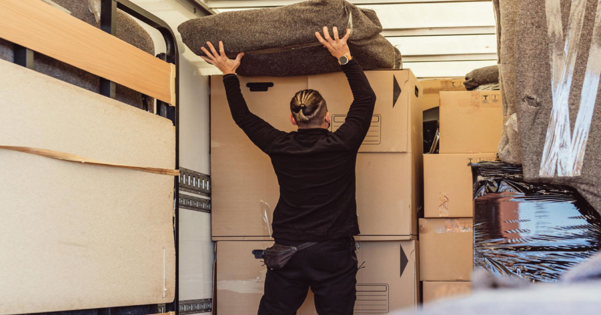 A person lifting a large blanket over a stack of cardboard boxes inside a moving truck.